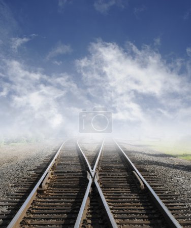 Two railroad tracks lead off into the daylight foggy sky with clouds.