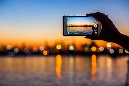 Tourist taking photo of sea harbor with mobile device at sunset
