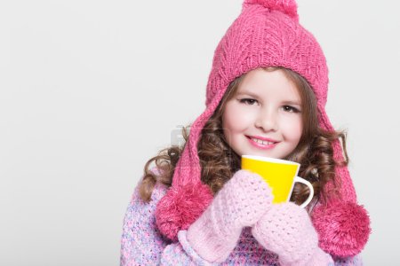 Beautiful child in winter hat drinking hot chocolate, little girl in woolen accessories with cup of tea or cocoa.