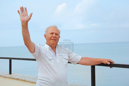 Smiling senior on veranda near seacoast, lifted hand upwards