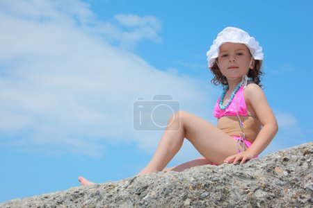 Little girl sits on rock against sky