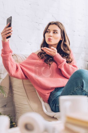 selective focus of cheerful girl sending air kiss while having video chat at smartphone