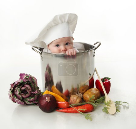 Portrait of a smiling baby sitting wearing a chef hat sitting inside a large cooking stock pot surrounded by vegetables and food, isolated on white