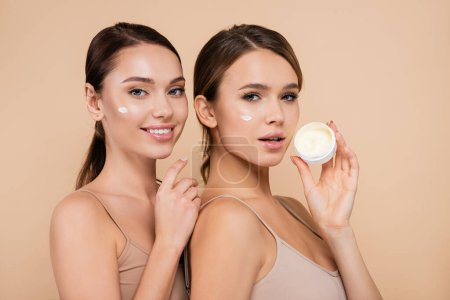 young woman holding cosmetic cream near smiling friend isolated on beige