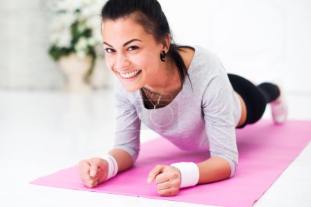 Pretty smiling young woman doing plank abdominal exercise at home in white room looking camera concept healthy lifestyle sport fitness