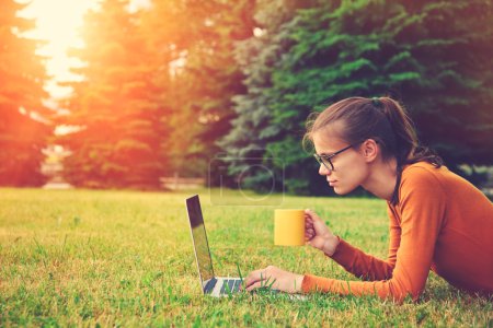 girl lying on the grass and using laptop and typing with coffee 