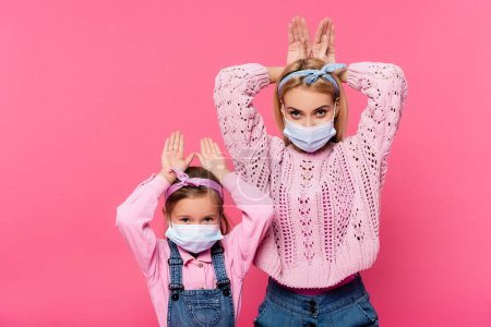 mother and daughter in medical masks making bunny ears with hands isolated on pink 