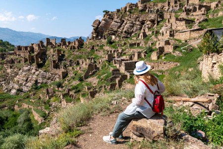 A young blonde girl in a hat, a traveler sits on a stone and looks at the village of Kahib Dagestan.