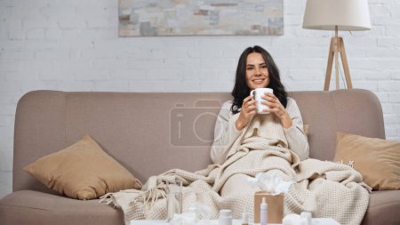 sick brunette woman holding cup of tea and smiling while sitting on couch