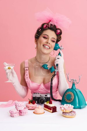 Young woman barbie in pink dress calling on landline phone while sitting at the table over isolated background