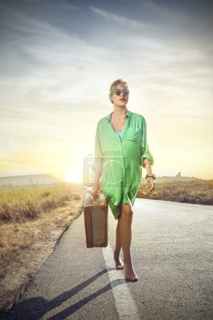 Beautiful woman carrying a suitcase while walking on a long sunny road