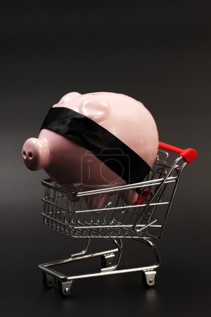 Shopping basket with pink piggy bank with black blindfold inside standing on black background - vertical