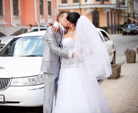 Portrait of bride and groom kissing in front of white limousine