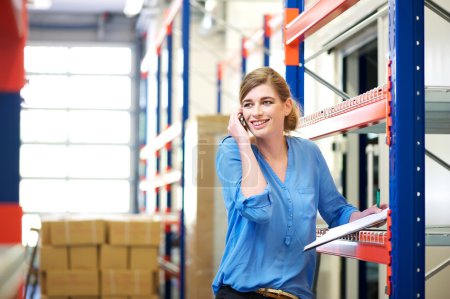 Female logistics worker controlling stock and talking on cellphone in warehouse