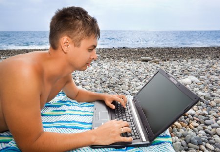Young man lying on stony beach, looking in the laptop screen