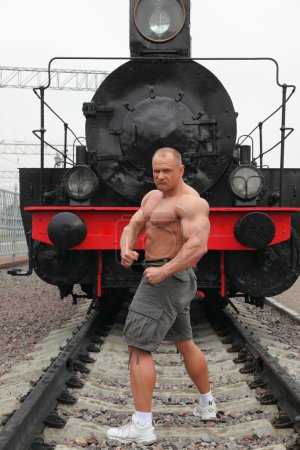 Strong shirtless man stands on railroad against locomotive