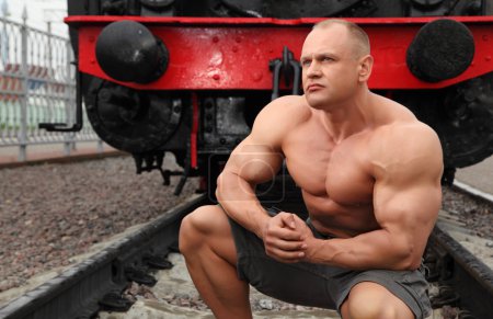 Strong shirtless man sits on railroad against locomotive
