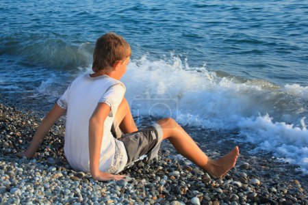 Sitting teenager boy on stone seacoast, wets feet in water, sitt