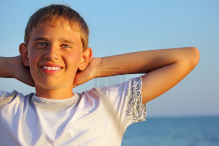Smiling teenager boy against sea, holding hands behind head