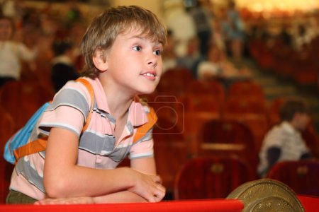 Little boy in circus leans on red rail looking into the distance