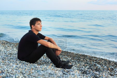 Sitting teenager boy on stone seacoast, Looking afar