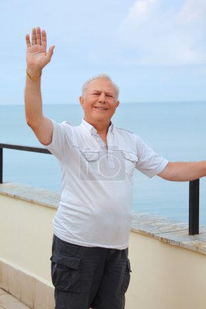 Smiling senior on veranda near seacoast, lifted hand upwards, ve
