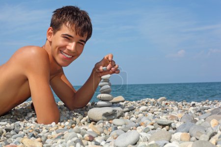 Teenager boy lying on stony seacoast, creates pyramid from pebbl