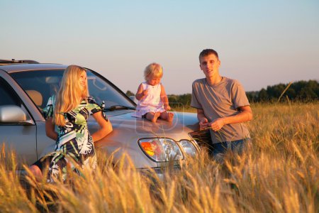 Parents and child sitting on car cowl on wheaten field