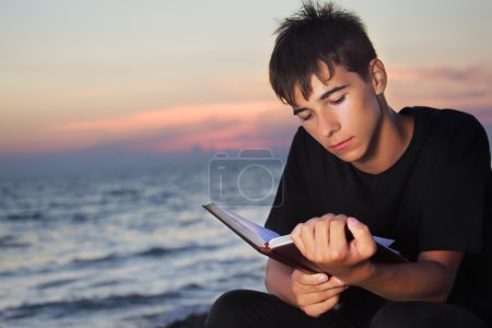 Teenager boy reads book sitting on beach in evening