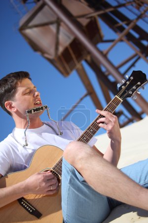 Guy plays guitar and lip accordion sitting on sand