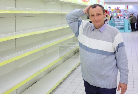 Elderly man at empty shelves in shop scratches in nape
