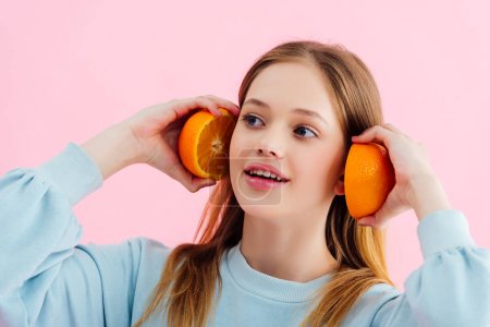 smiling pretty teenage girl holding orange halves near ears isolated on pink