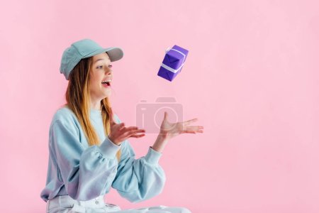 excited pretty teenage girl in cap throwing gift in air isolated on pink