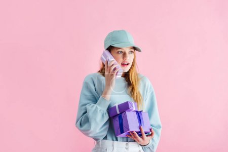 surprised pretty teenage girl in cap holding present as telephone isolated on pink