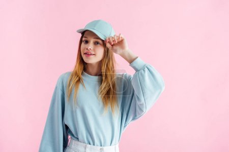smiling pretty teenage girl touching cap isolated on pink
