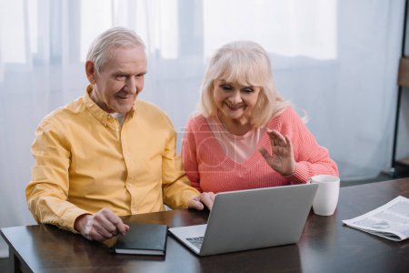 senior couple sitting at table and using laptop during video call at home