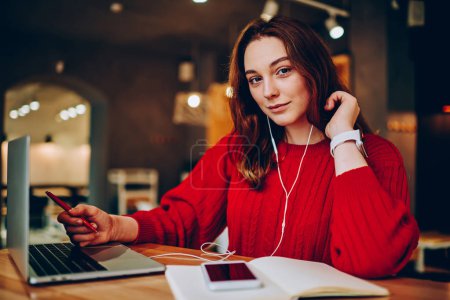 Portrait of smart female student using modern technology for learning language on her own, teen girl looking at camera listening record via cellular and earphones preparing homework in coworking offic
