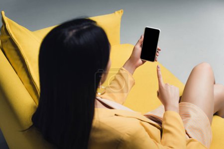 brunette businesswoman sitting on yellow sofa and pointing with finger at smartphone with blank screen isolated on grey