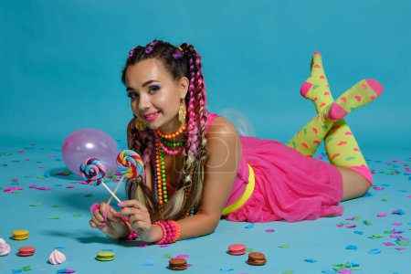 Lovely girl with a multi-colored braids hairstyle and bright make-up, posing in studio with lollipop, air balloons and confetti against a blue background.