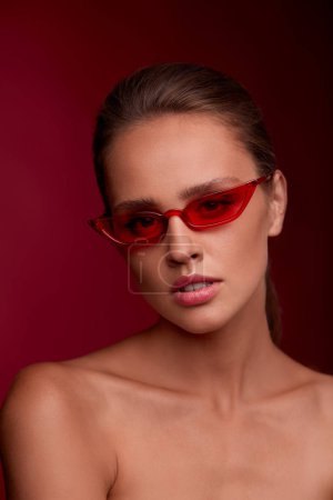 Portrait of beautiful young woman with perfect makeup wearing red sunglasses. Smiling fashion model in cat eye sunglasses posing on burgundy background. Studio shot. Summer vacation.