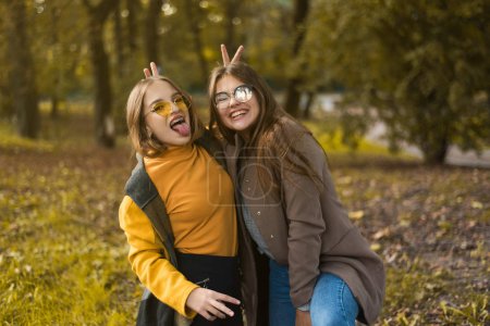 Two teenage girls are having fun in the park, autumn outfit. Funny girl friends throwing dry leaves in the city in autumn. Happy family on autumn walk! Geasture peace.
