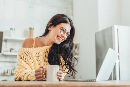 young laughing female freelancer sitting at wooden table with laptop and coffee cup in kitchen at home