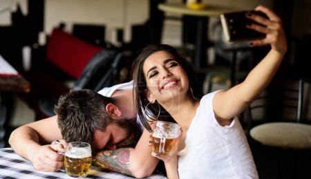 Girl taking selfie photo drunk boyfriend. Take selfie to remember great event. He appears too weak for her. Woman making fun of drunk friend. Man drunk fall asleep table and girl with full beer glass