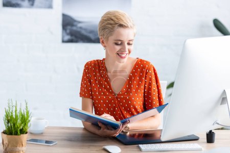 smiling female freelancer reading book at table with graphic tablet and computer in home office 