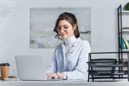 Smiling businesswoman in glasses typing on laptop at workplace