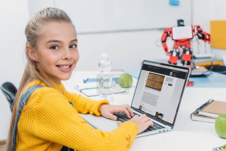 smiling schoolgirl sitting at table with robot model, looking at camera and using laptop with science website on screen during STEM lesson  