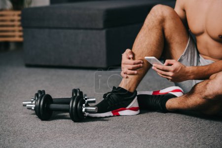 cropped view of man sitting on floor near dumbbells and using smartphone 