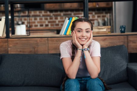 smiling teenage girl with hands on cheeks looking at camera and sitting on sofa at home