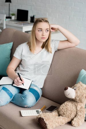 thoughtful teen student girl with notebook sitting on couch and looking away