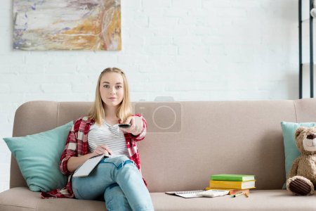 smiling teen student girl with remote control watching tv while sitting on couch with book and notes